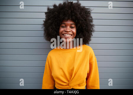 Young black woman with afro standing against grey security shutters, smiling to camera, close up
