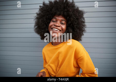 Young black woman with afro standing against grey security shutters, laughing to camera, close up