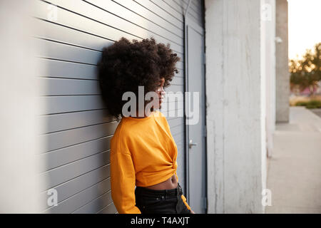 Young black woman with afro leaning in the street against grey security shutters, side view