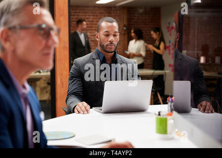 Businessman Working At Desk On Laptop In Open Plan Office With Colleagues In Background Stock Photo