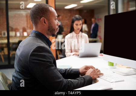 Businessman Working At Desk On Computer In Open Plan Office With Colleagues In Background Stock Photo