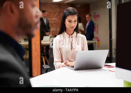 Businesswoman Working At Desk On Laptop In Open Plan Office With Colleagues In Background Stock Photo