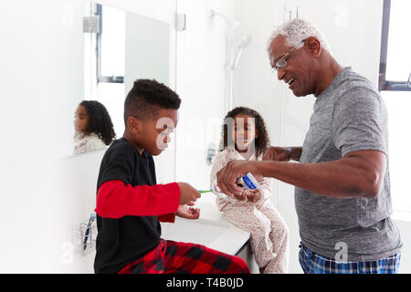 Grandfather In Bathroom Wearing Pajamas Helping Grandchildren To Brush Teeth Stock Photo