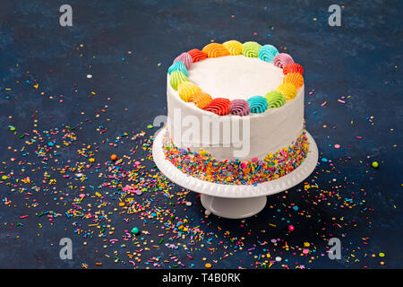 White Birthday cake with rainbow icing and colorful Sprinkles over a dark blue background. Stock Photo