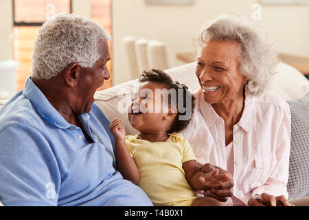 Grandparents Sitting On Sofa At Home Playing With Baby Granddaughter Stock Photo
