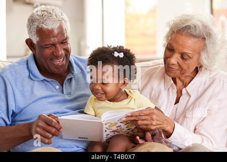 Grandparents Sitting On Sofa At Home Reading Book With Baby Granddaughter Stock Photo