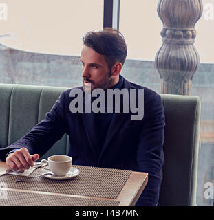 Thinking handsome bearded man sitting in cafe and drinking cup of coffee on the breakfast. Closeup portrait Stock Photo