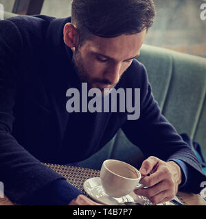 Thinking handsome bearded man sitting in cafe and drinking cup of coffee on the breakfast. Closeup toned color portrait Stock Photo