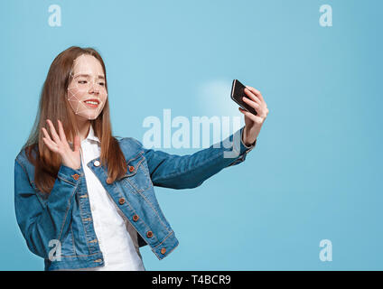 Trying something new. A girl with a smartphone in jeans suit standing on blue background. Technology of face recognition on polygonal grid. Concept of cyber security, business, work, education. Stock Photo