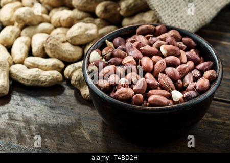 peeled peanuts in a plate and unpeeled one scattered on a brown wooden table. space for text Stock Photo