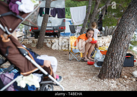 Mother with children at the campsite, preparing food in camp kitchen, checking on sleeping baby in a stroller. Active natural lifestyle, family time,  Stock Photo