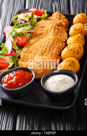 Delicious fried fillet of white fish in breading with a side dish of new potatoes and fresh vegetable salad close-up on a plate on the table. vertical Stock Photo