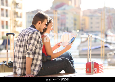 Two happy tourists checking paper map and smart phone on holiday in a coast town Stock Photo