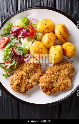 Serve fried steaks in sesame breading with new potatoes and fresh salad close-up on a plate on the table. Vertical top view from above Stock Photo