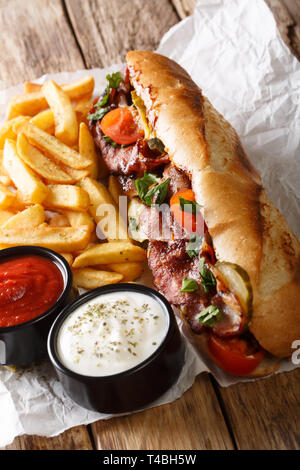 Delicious hot dog with bacon and vegetables served with french fries and sauces close-up on the table. vertical Stock Photo
