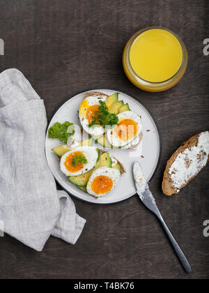 Toast with cream cheese, avocado and eggs on white plate with knife and napkin at wooden background Stock Photo