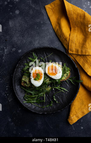 Tasty boiled egg on arugula salad with grain bread on black plate with orange napkin at black background. Concept of healthy food. Flat lay, table top Stock Photo