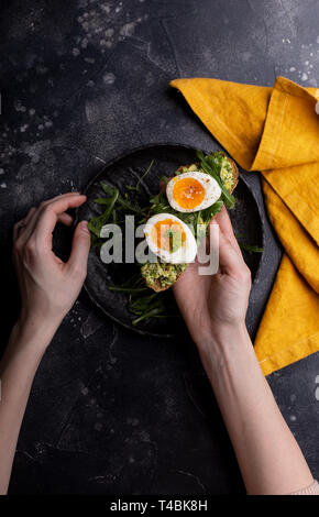 Sandwich with egg and avocado on bread in woman's hands on black background with orange napkin. Table top, flat lay. Concept of healthy breakfast. Stock Photo