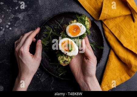 Toast with egg and avocado on bread in women's hands on black background. Top view, table top, flat lay. Concept of healthy breakfast. Lifestyle photo Stock Photo