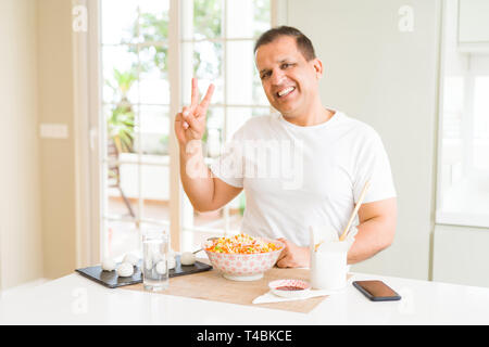 Middle age man eating asian food with chopsticks at home smiling with happy face winking at the camera doing victory sign. Number two. Stock Photo