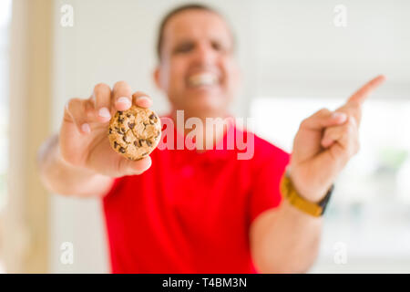 Middle age man eating chocolate chips cookies at home very happy pointing with hand and finger to the side Stock Photo