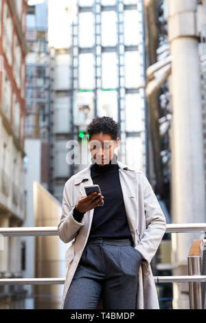 Fashionable young black woman standing in the city leaning on a hand rail using her smartphone, vertical Stock Photo