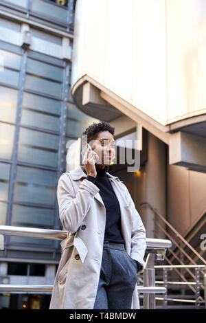 Fashionable young black businesswoman standing against handrail in the city talking on smartphone, vertical Stock Photo