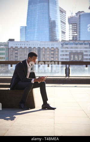 Millennial black businessman wearing black suit and white shirt sitting on the Thames embankment, London, using smartphone, vertical Stock Photo