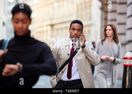 Millennial black businessman walking in a busy London street using smartphone, selective focus Stock Photo