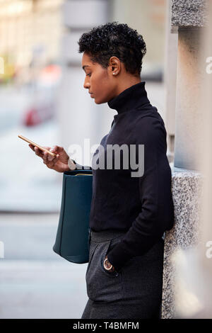Young black woman standing on a street in London using her smartphone, side view, close up, vertical Stock Photo