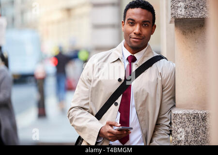 Young black businessman leaning on a wall in the street in London holding phone and smiling to camera, selective focus Stock Photo