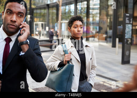 Two business people walking in a street in the city of London, man using smartphone and woman carrying a bag, selective focus Stock Photo