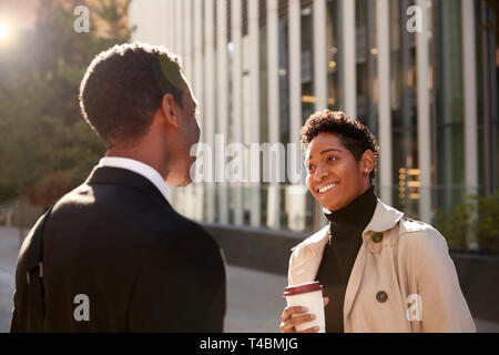 Smiling young black businesswoman standing on the street with a takeaway coffee, talking to her male colleague, selective focus Stock Photo