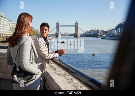 Two young adult women standing at the Thames riverside talking, Tower Bridge in the background Stock Photo