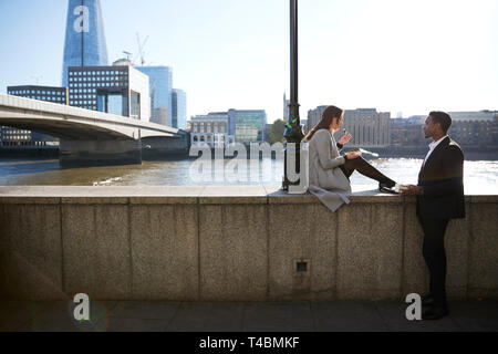 Two millennial colleagues take a break sitting on the embankment eating near London Bridge by the River Thames, backlit Stock Photo