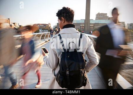 Back view of young adult woman standing on Millennium Bridge, London, using smartphone, motion blur Stock Photo