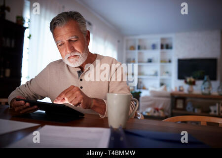 Senior Hispanic man sitting at his dining table reading an e book at home in the evening, close up Stock Photo