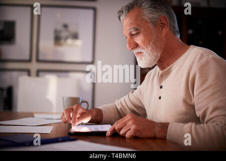 Senior Hispanic man sitting at a table using a tablet computer  in the evening, close up, side view Stock Photo