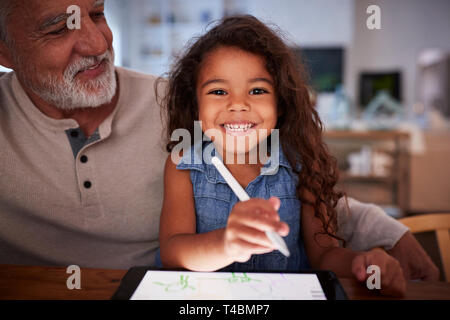 Senior Hispanic man with his young granddaughter using stylus and tablet computer, smiling to camera Stock Photo