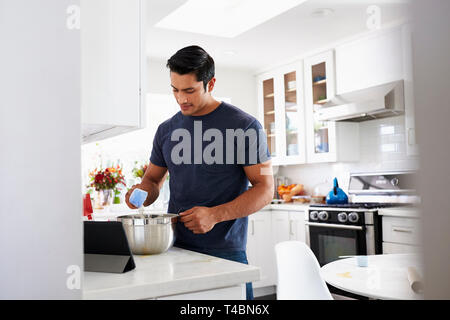 Millennial Hispanic man preparing cake mixture in kitchen, following a recipe on a tablet computer