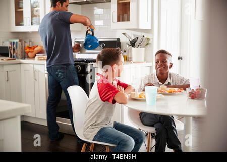 Two pre-teen male friends sit talking in kitchen at one boy’s house, dad in the background Stock Photo