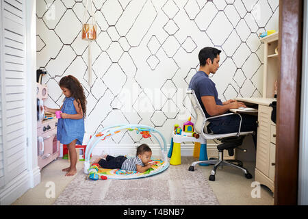 Dad sits working at a desk at home while his baby son and young daughter play in the room behind him Stock Photo
