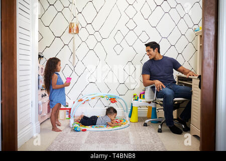 Dad working at a desk at home turns around to talk to his young kids, playing in the room behind him Stock Photo