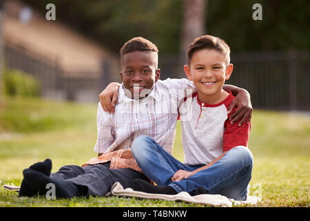 Two pre-teen male friends sitting on the grass in a park, arms around each other, looking to camera Stock Photo