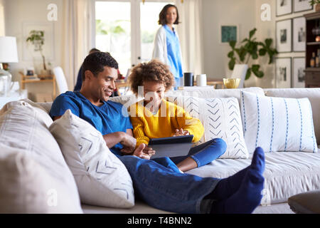 Close up of young father sitting on sofa in the living room with his pre-teen daughter using tablet computer, selective focus, mum in the background