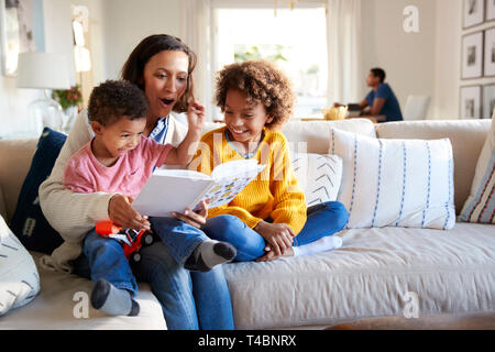 Close up of young mother sitting on a sofa in the living room reading a book to her two children, father sitting at a table in the background, focus on foreground
