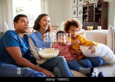 Young family sitting together on the sofa in their living room watching TV and eating popcorn Stock Photo