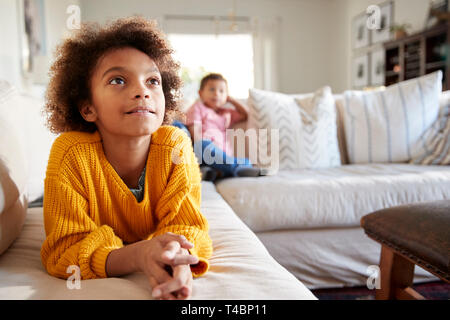 Close up of pre-teen girl lying on sofa watching TV in the living room, her younger brother sitting in the background, focus on foreground Stock Photo