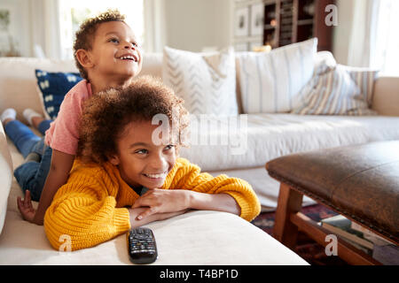 Pre-teen girl lying on sofa watching TV in the living room with her younger brother sitting on her back, close up Stock Photo
