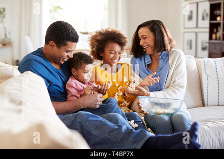 Young mixed race family sitting together on the sofa in their living room watching a scary movie, clearing up spilled popcorn, selective focus Stock Photo